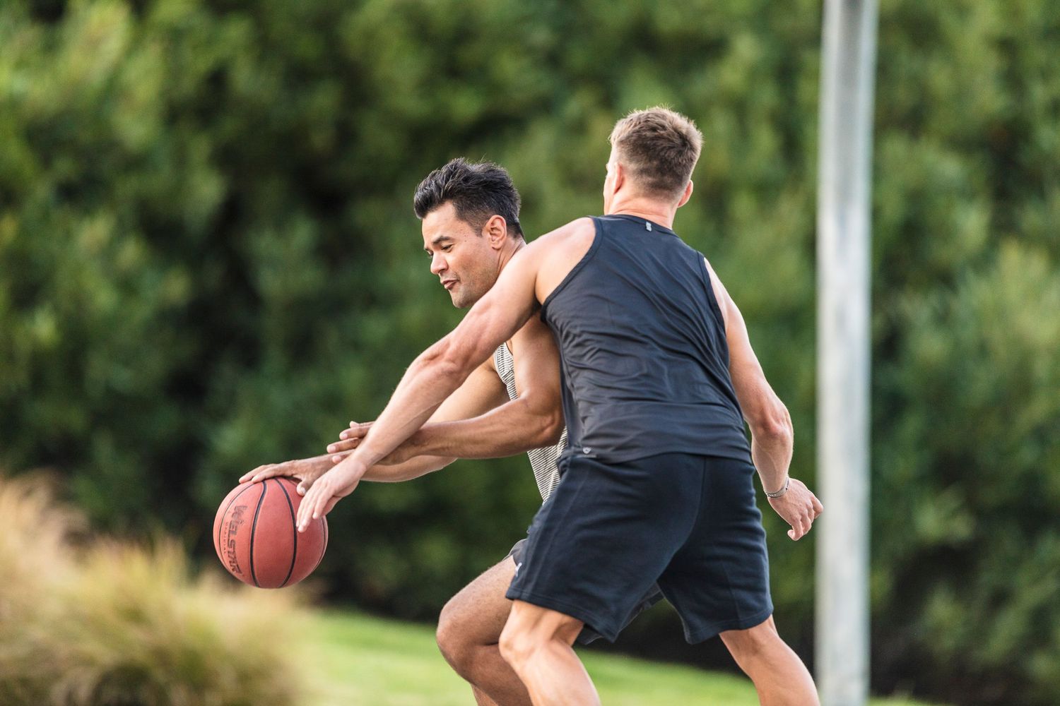 Two athletes playing basketball outdoors, one defending against the other's dribble.