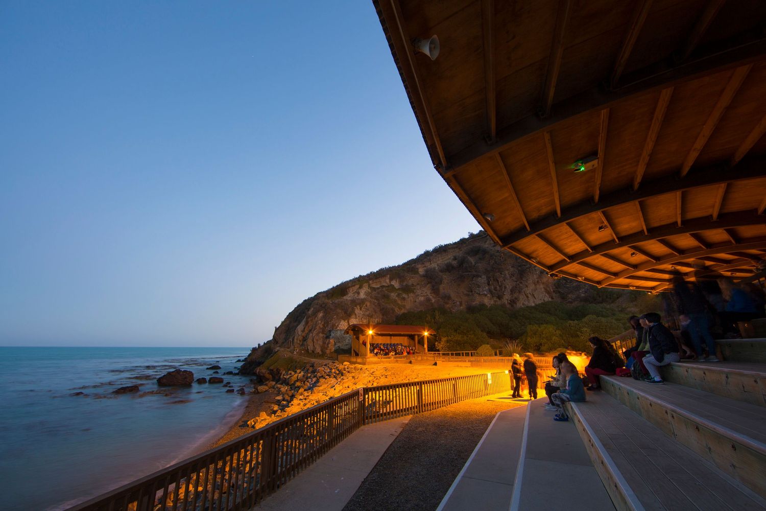 Scenic coastal pathway lit by warm lights at dusk with ocean views and rocky cliffs.