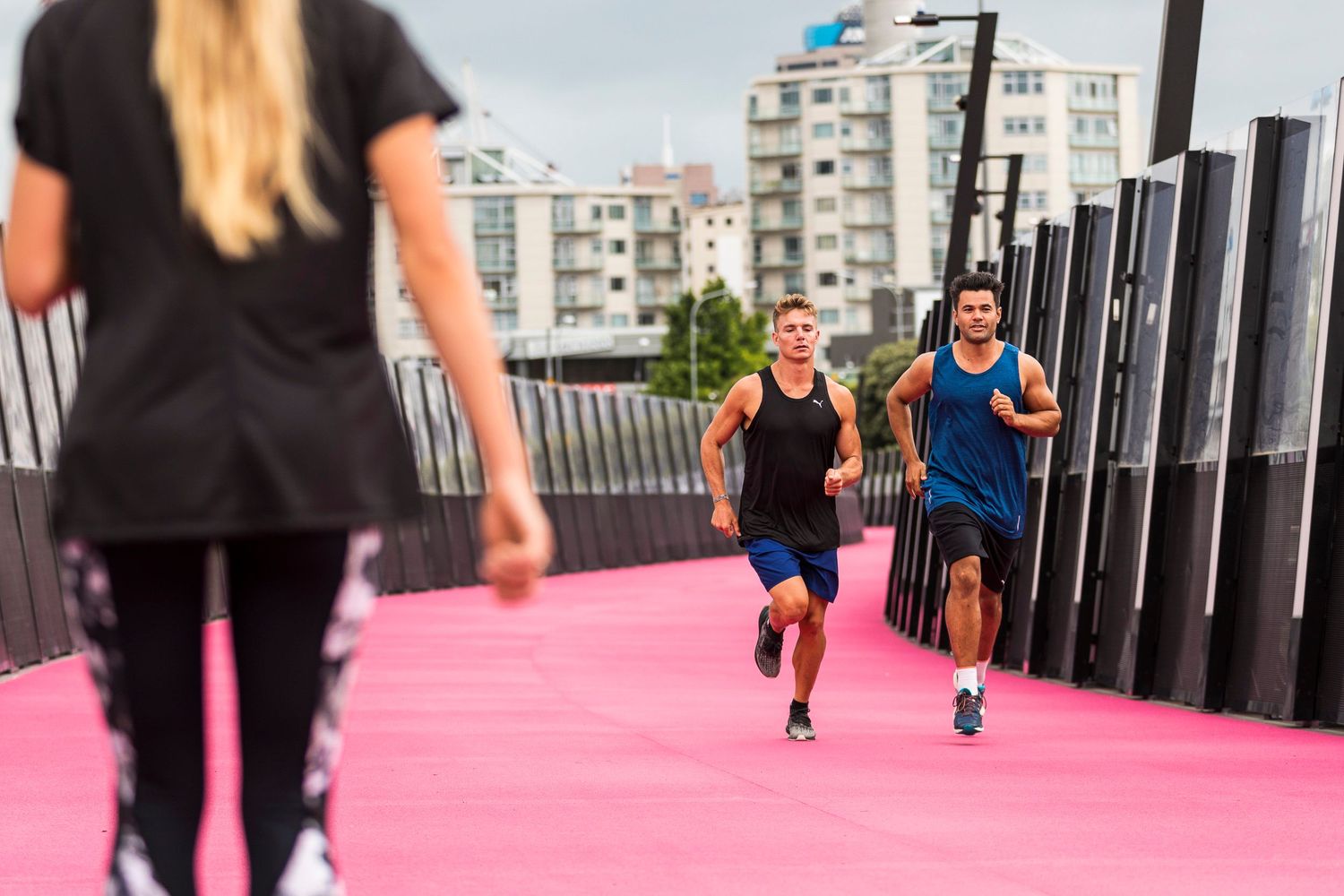 Two people running on a bright pink outdoor track with urban buildings in the background.