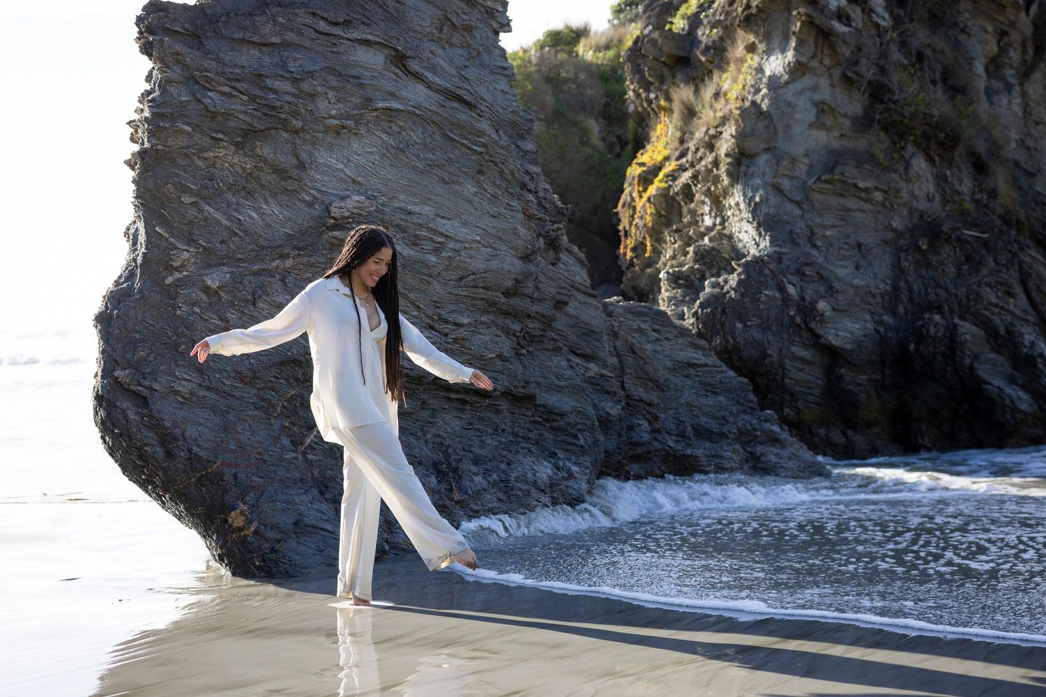 Figure in white clothing dancing on a beach near dramatic rocky cliffs.