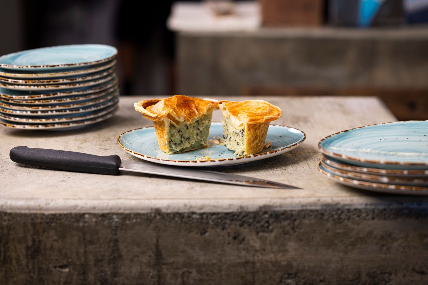 Sliced savory paua pie served on blue-rimmed plates with a knife on a concrete counter.