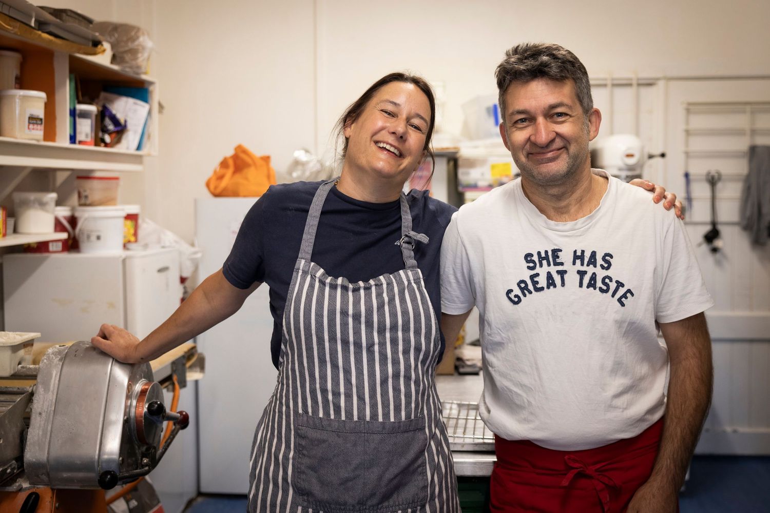 Two chefs posing together in a kitchen, one wearing a striped apron and one wearing a graphic t-shirt.