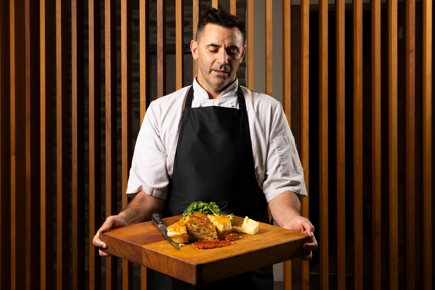 Chef in black apron presents plated dish on wooden board against slatted wall.