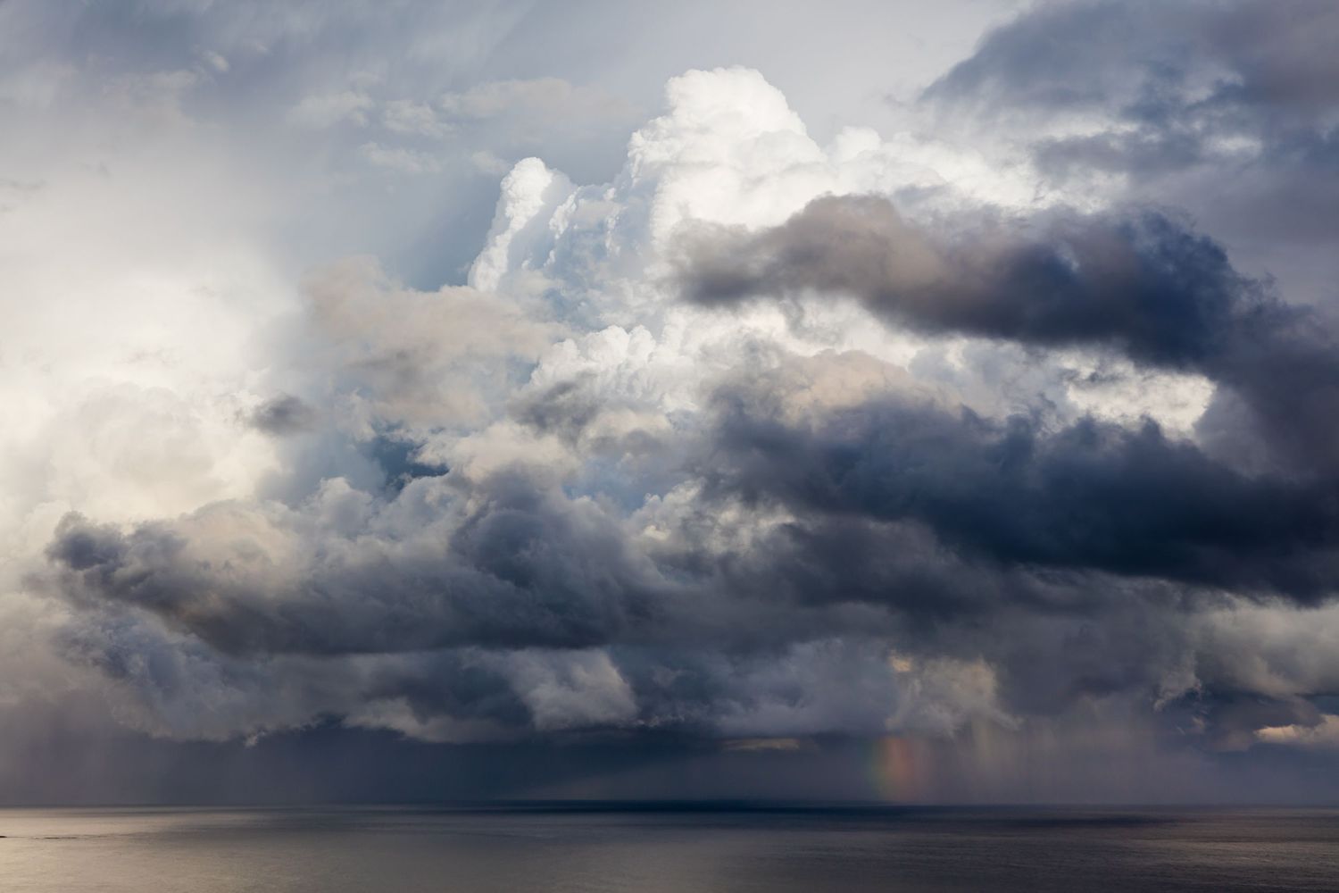 Dramatic storm clouds gather over ocean horizon with hints of rainbow peeking through.