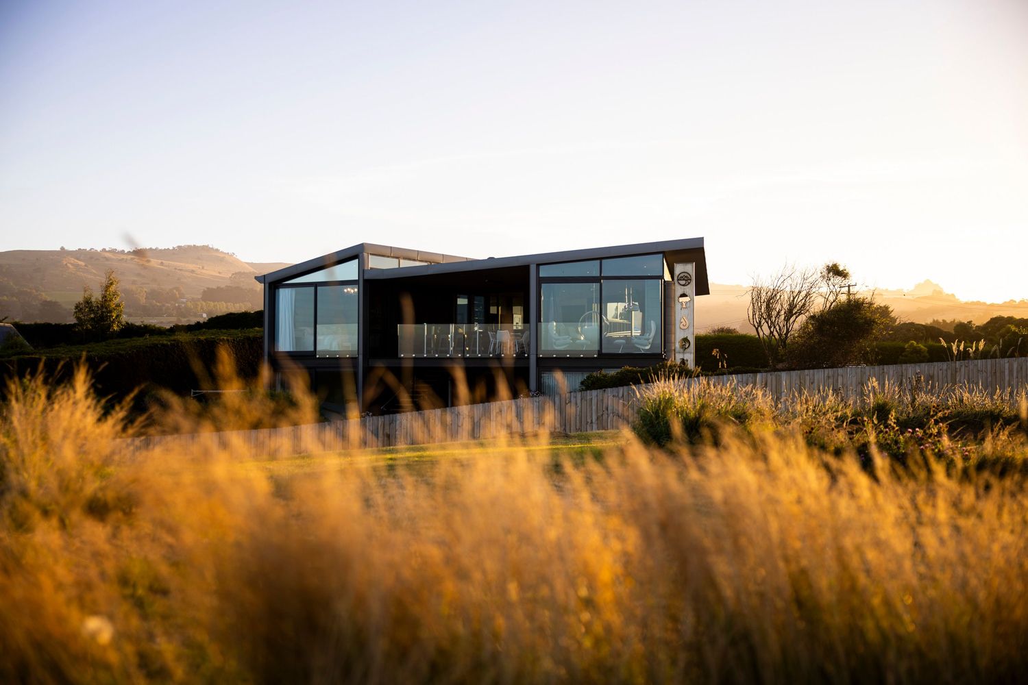 Modern home with floor-to-ceiling windows viewed through swaying grass at sunset.