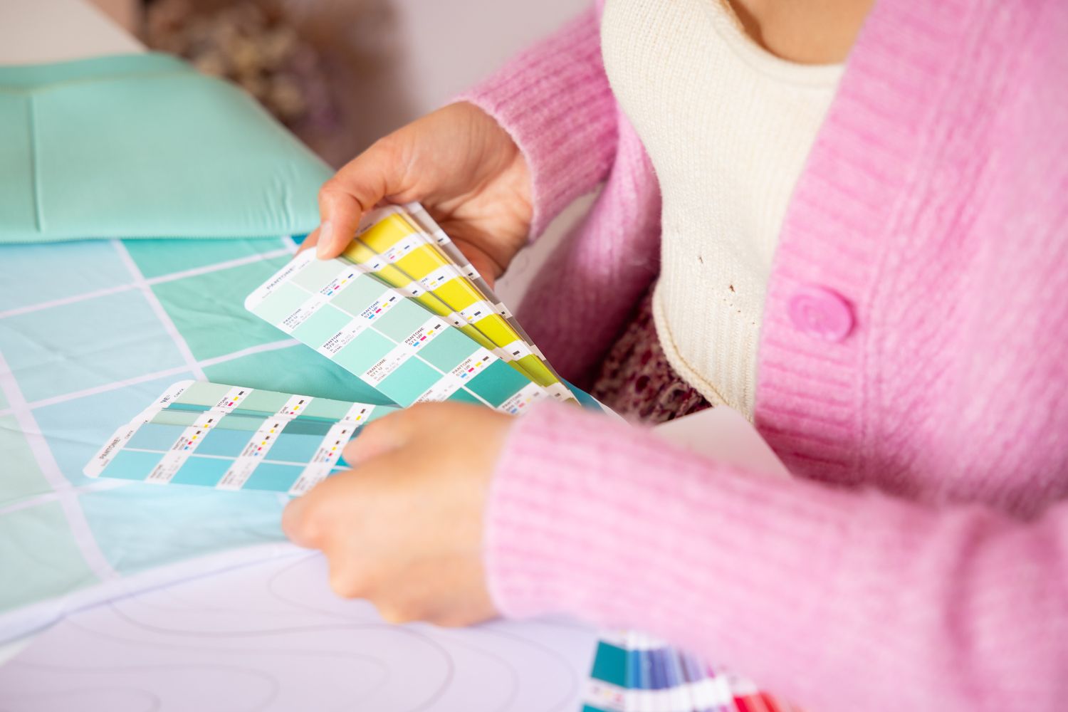 Hands holding paint color swatches over a desk with mint green and pink accessories.