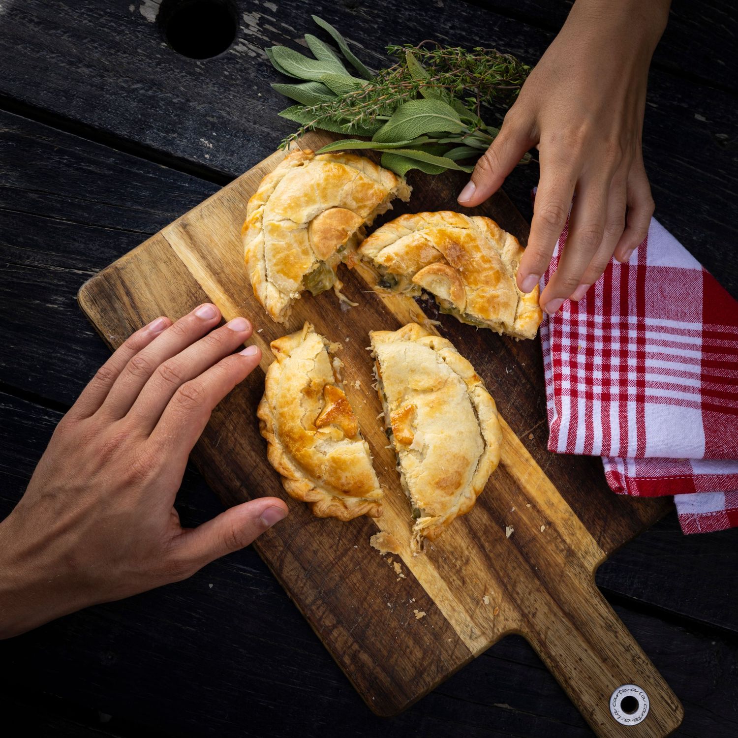 Hands reaching for golden-brown pastries on a wooden cutting board with red checkered napkin.