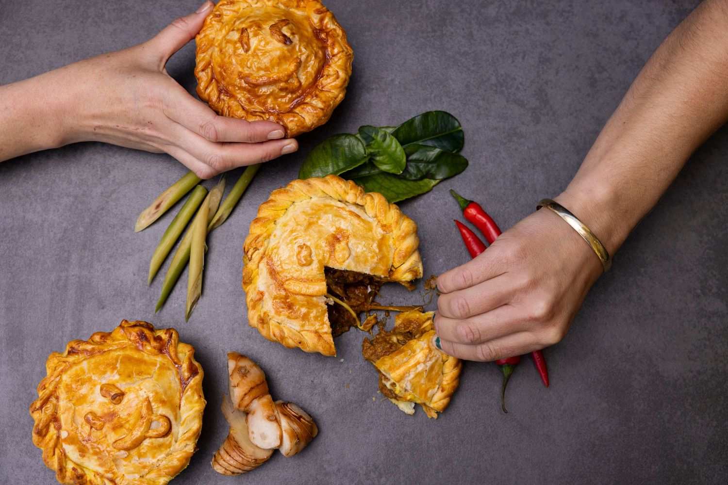 Hands breaking open fresh baked pies with galangal and herbs on dark surface.