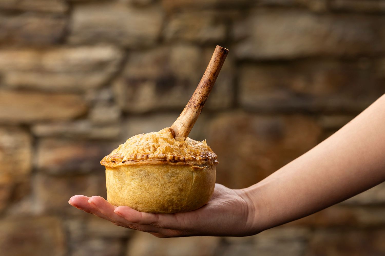 Hand holding rustic pot pie with bone handle against stone wall background.