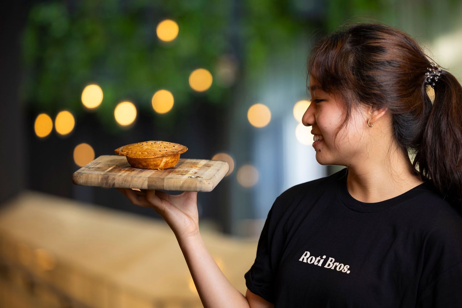 Server in black uniform holds up a wooden plate with food in front of decorative lighting.
