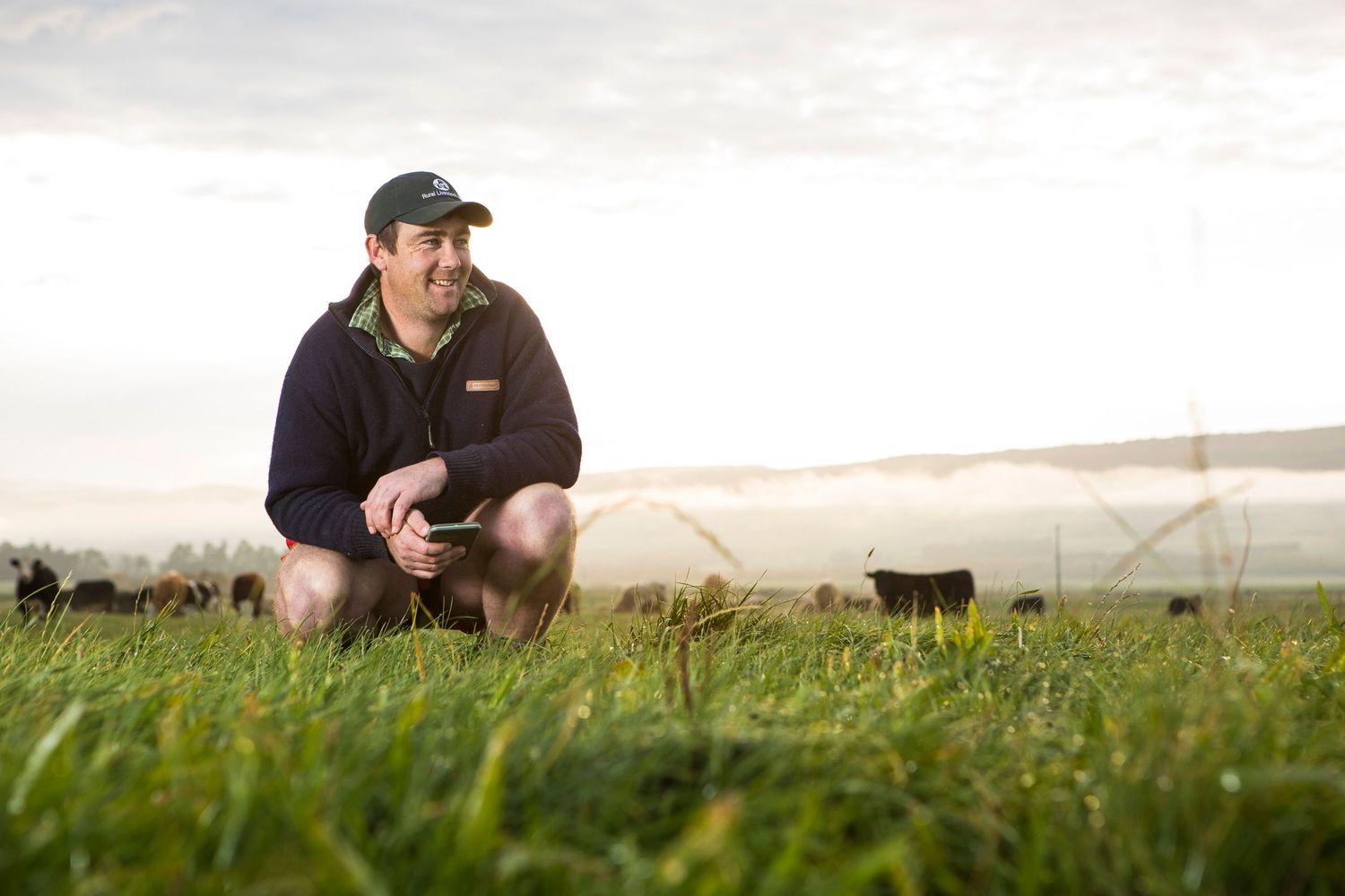 Farmer kneeling in green pasture at sunset with irrigation system in background.