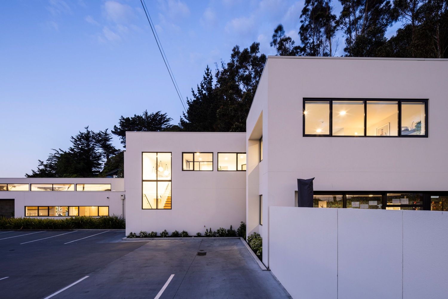Illuminated modern building exterior at dusk with large windows and clean white walls.
