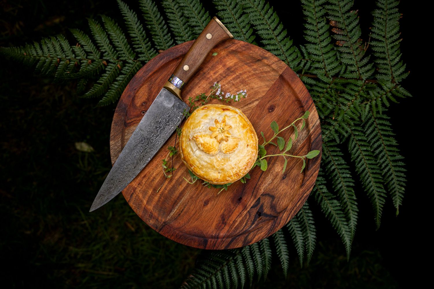 Golden pie on wooden cutting board with knife and fern leaves.