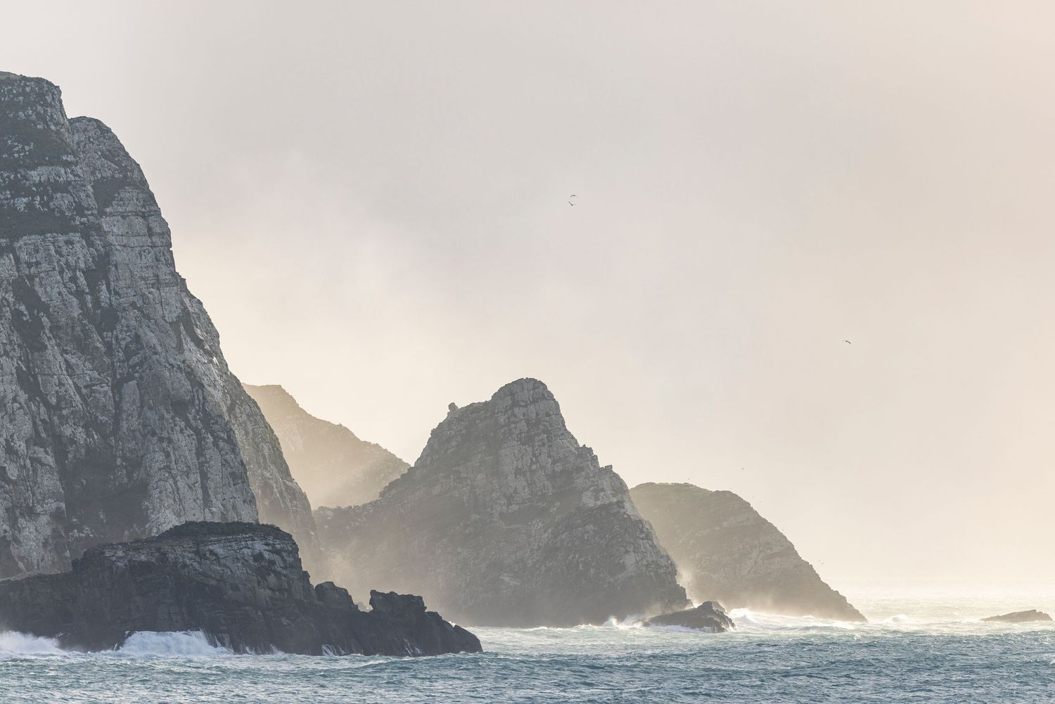 Misty coastal cliffs and rock formations emerging from the sea under a moody sky.