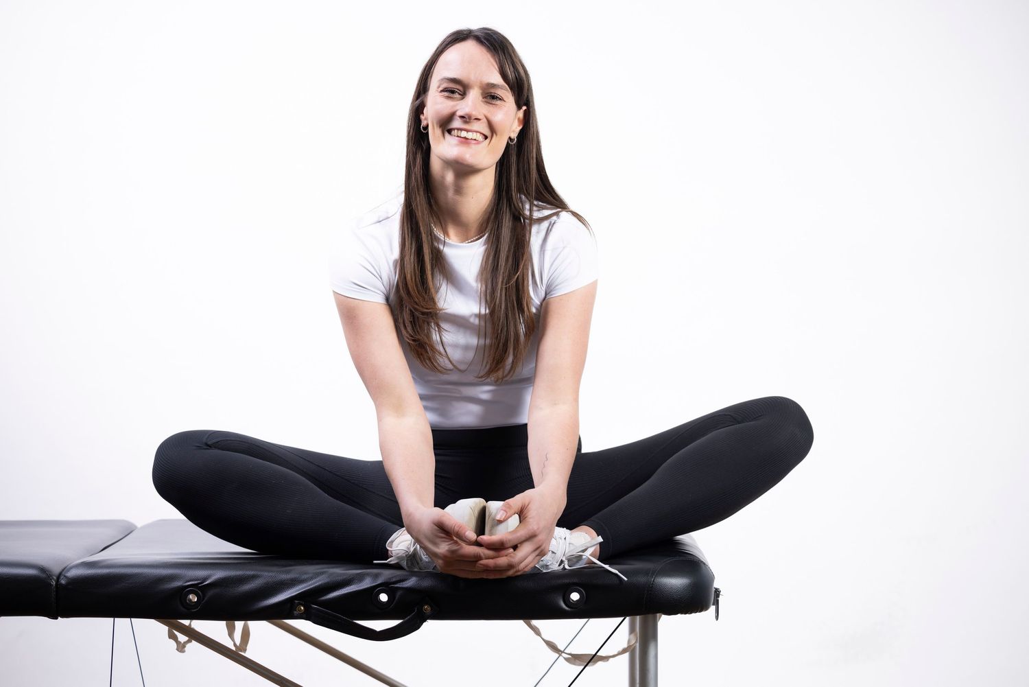 Person sitting cross-legged in a relaxed pose on a massage table wearing white shirt and black pants.
