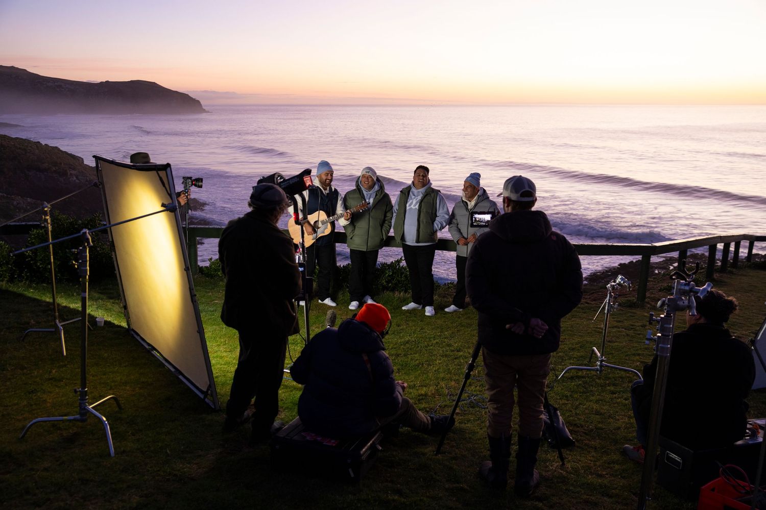 Film crew setting up equipment on coastal cliff during sunset shoot.