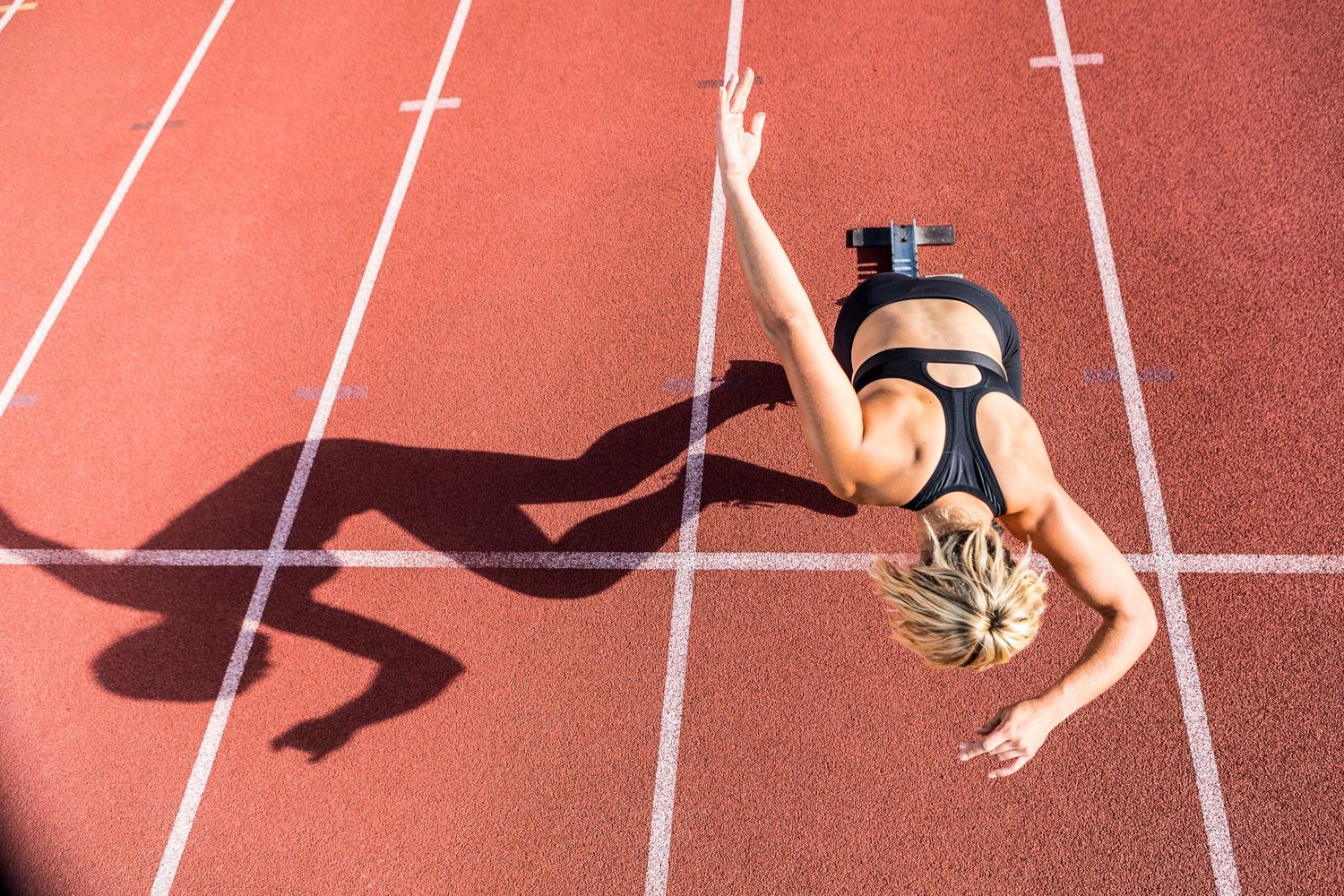 An athlete stretches on a red running track creating a dynamic shadow in sunlight.