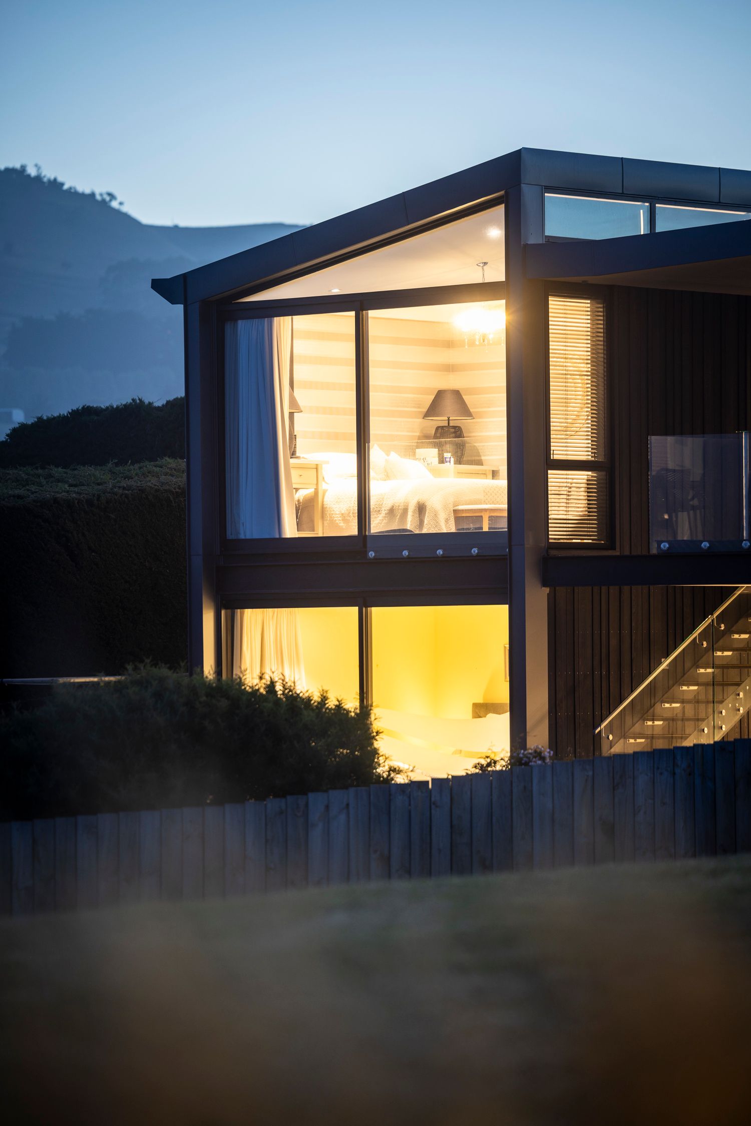 Modern mountain house with illuminated glass windows glows warmly at dusk against a mountainous backdrop.