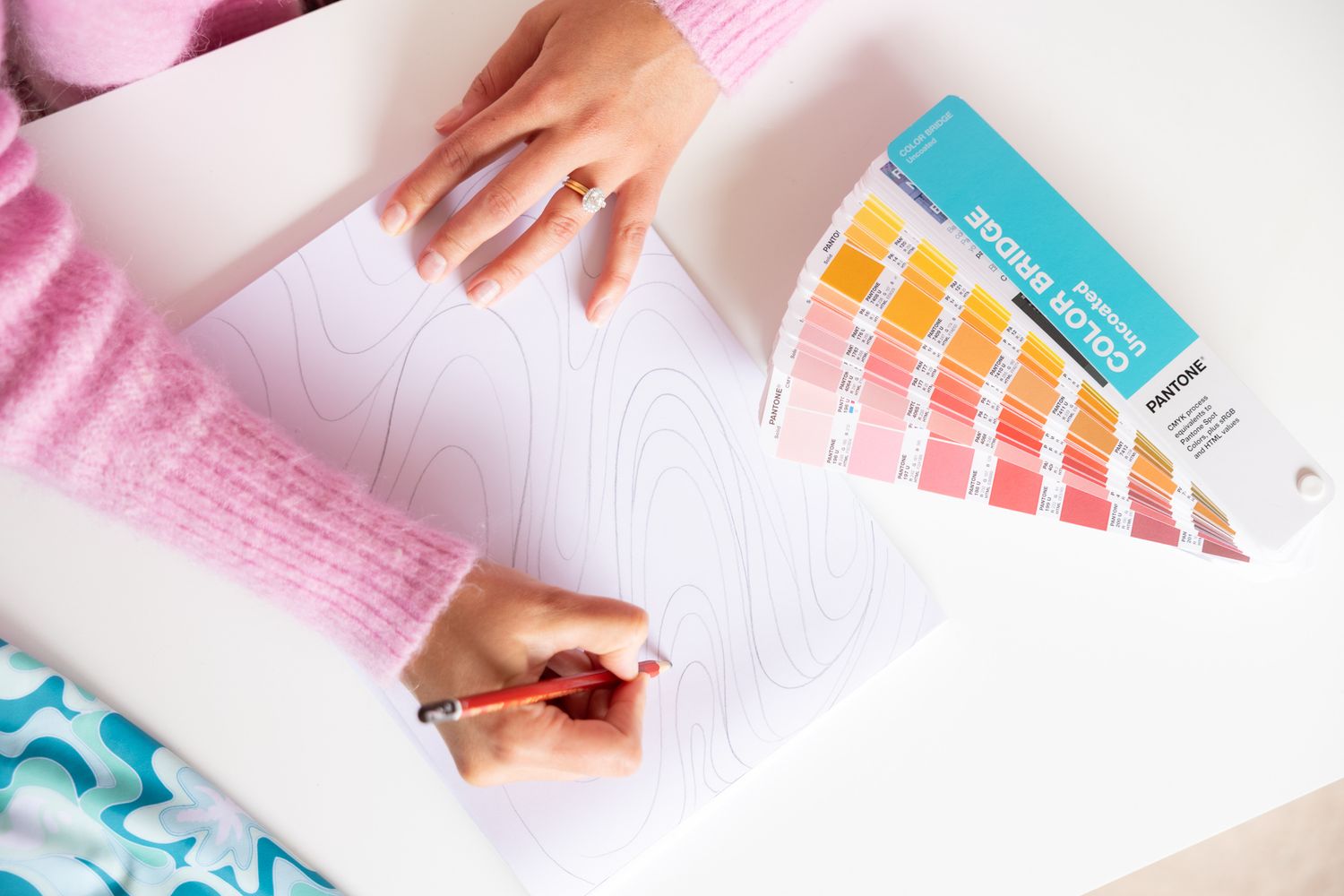 Hands holding color swatches and drawing on white paper with pink sweater sleeves visible.