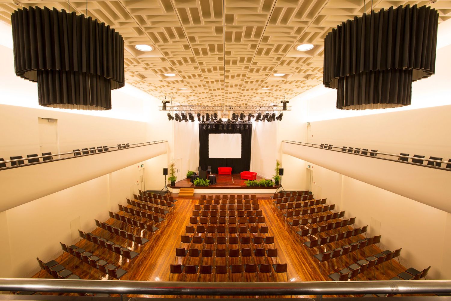 Concert hall with acoustic panels on ceiling and wooden seats arranged in symmetrical rows facing stage.