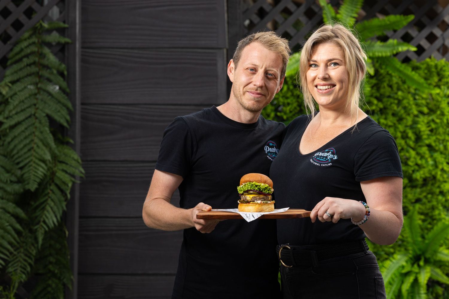 Staff members in black uniforms present a gourmet burger against a lush green garden backdrop.