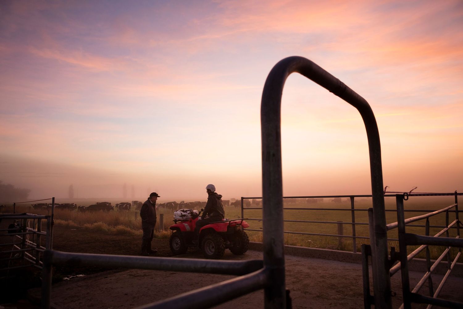 Silhouette of ATV rider against dramatic sunset sky with metal railing in foreground.