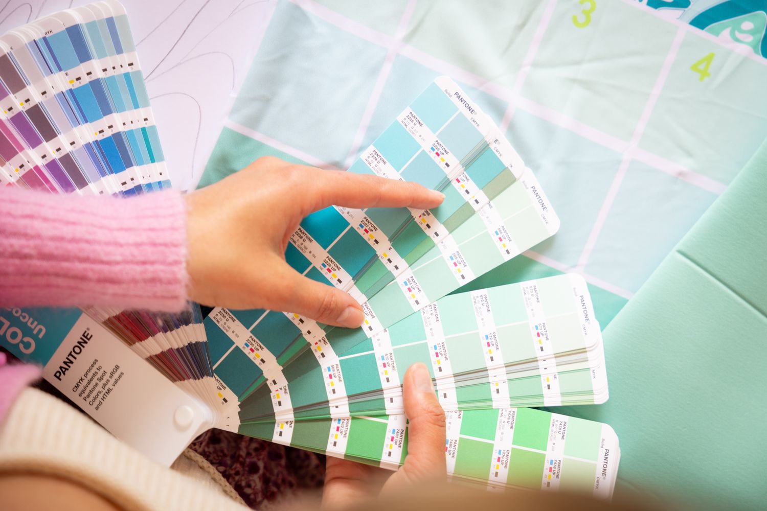 Hands selecting colors from paint swatches and design samples in pastel green and blue tones.
