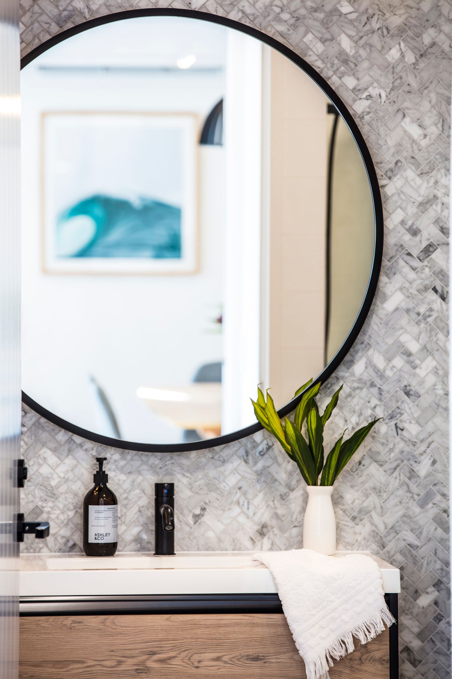 Modern bathroom featuring round mirror, marble wall, and coastal artwork reflection.
