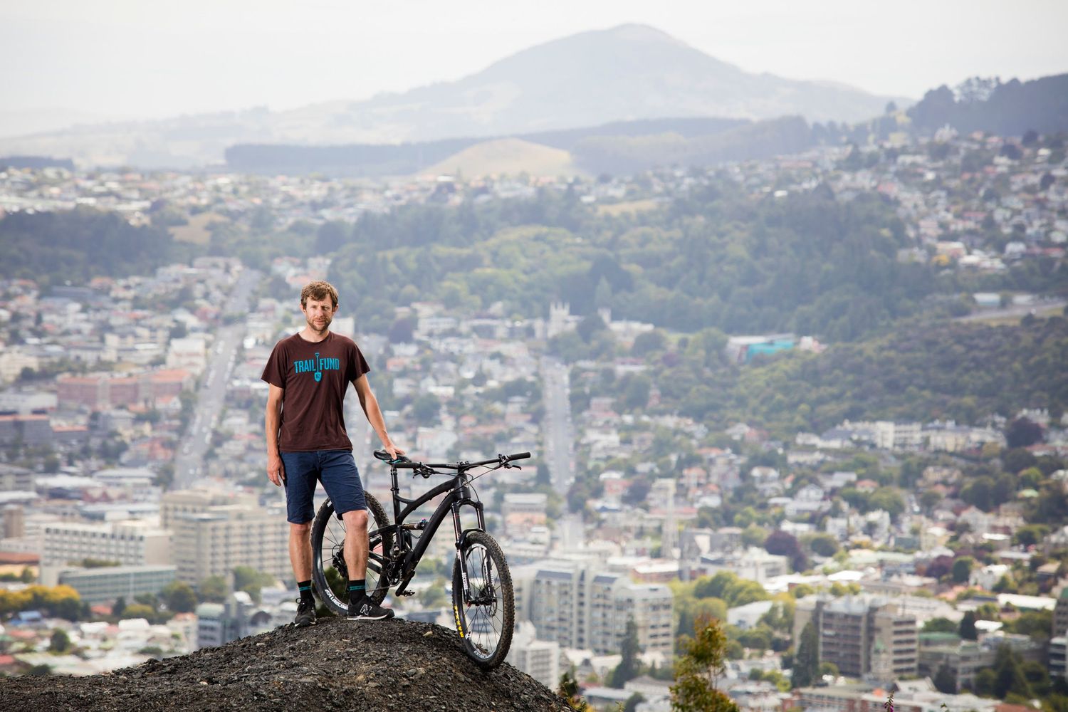 Mountain biker stands with bicycle overlooking cityscape from elevated viewpoint.