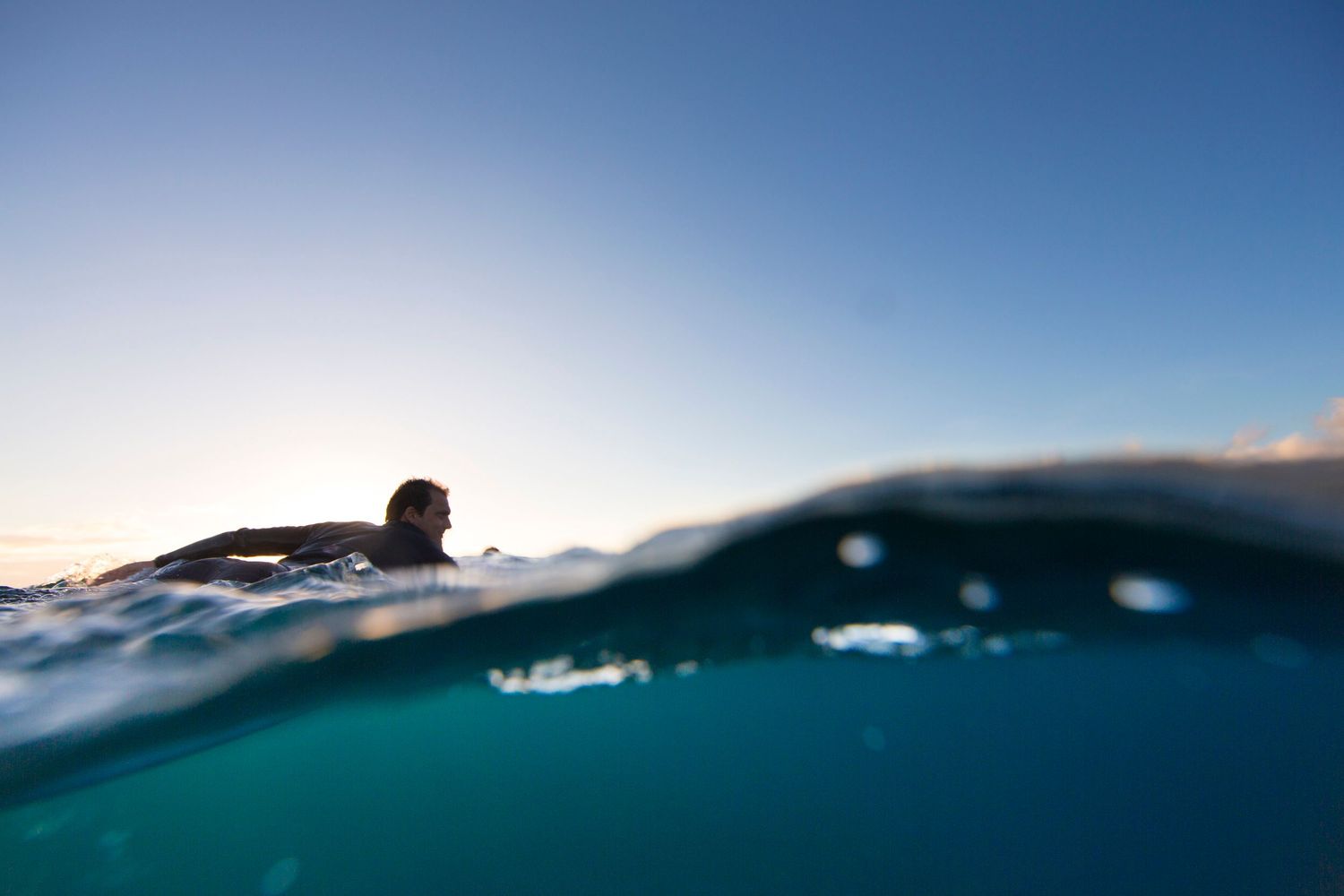 Minimalist split-shot photograph of swimmer in turquoise ocean water against bright sky.