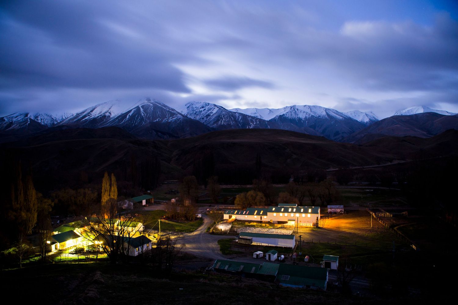 Nighttime view of illuminated mountain village with snow-capped peaks in background.