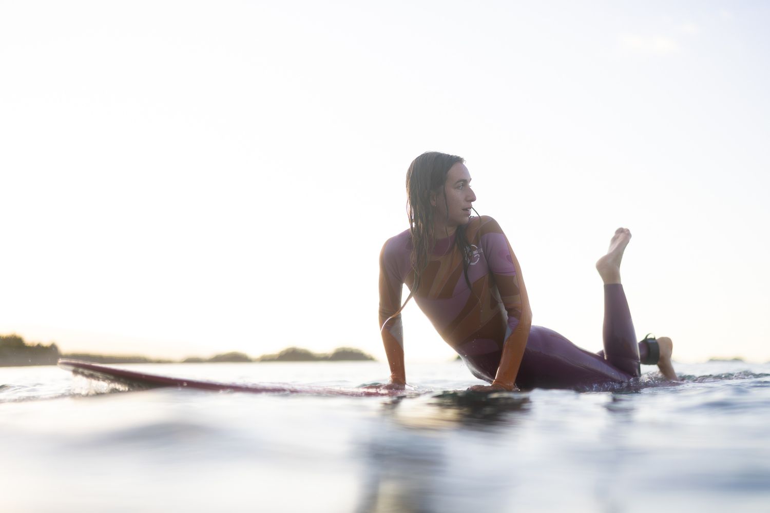 A surfer lies on a surfboard in calm ocean waters during sunset.