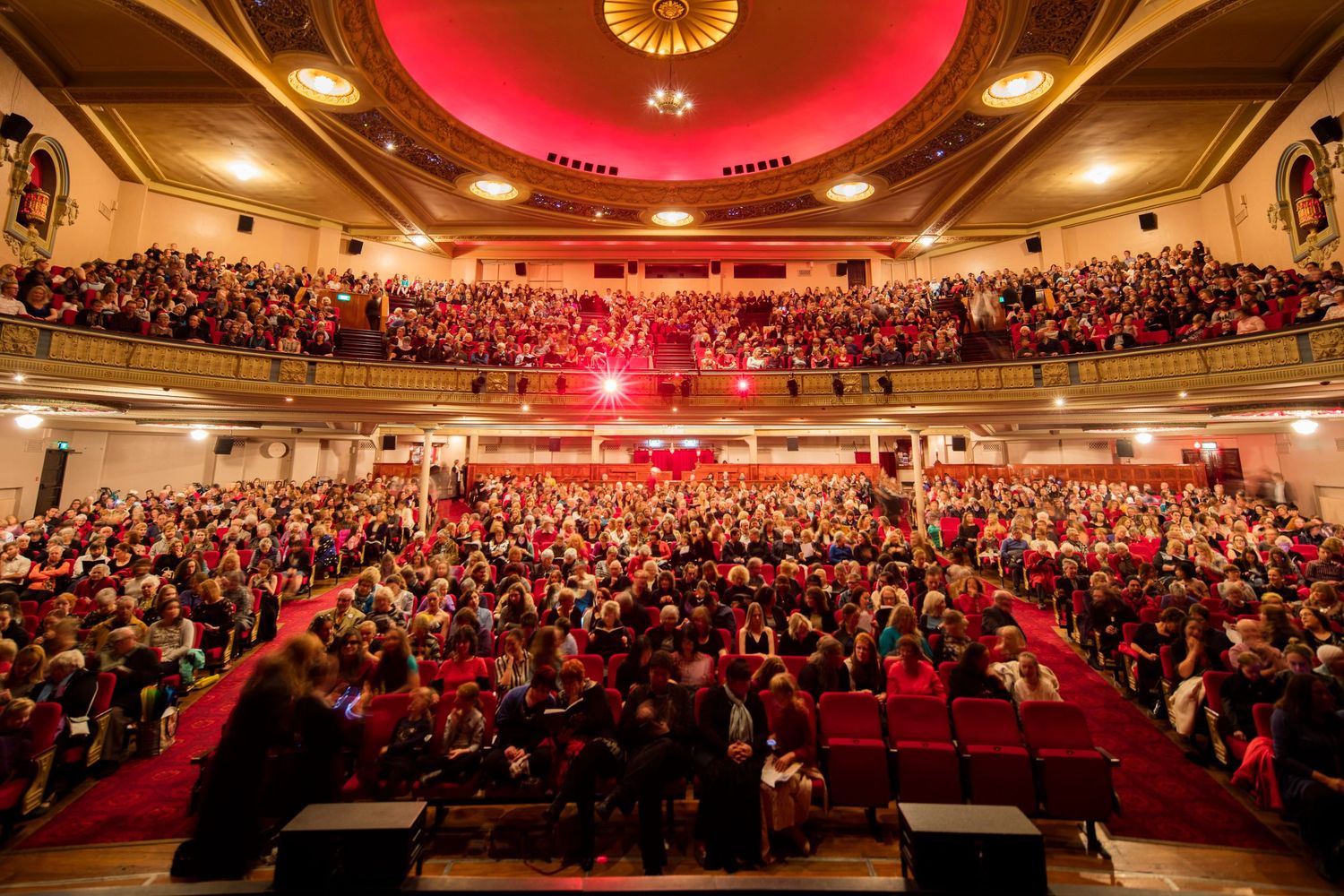 Panoramic view of packed concert hall with red ceiling lights and ornate architectural details.