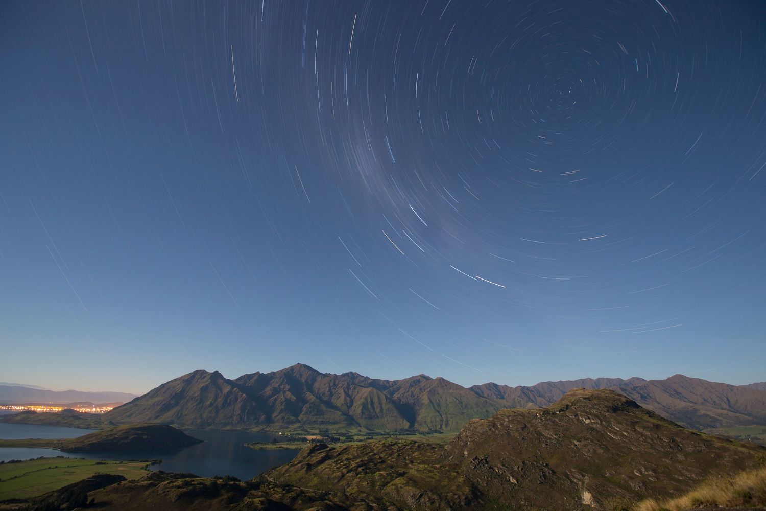Star trails streak across the night sky above rugged mountain peaks and a tranquil lake below.