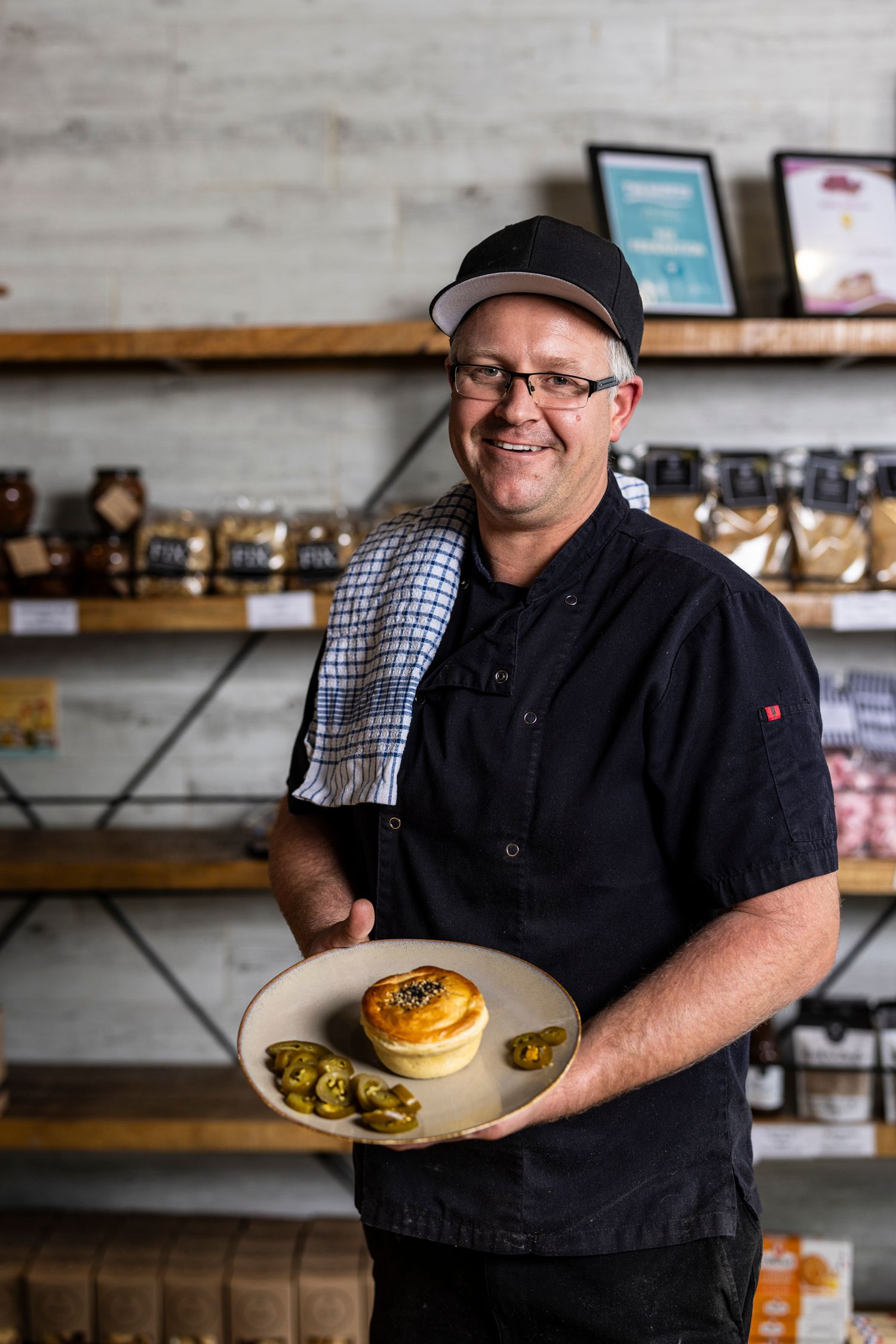 Chef in black uniform presenting a plate of food in front of shelved retail products.