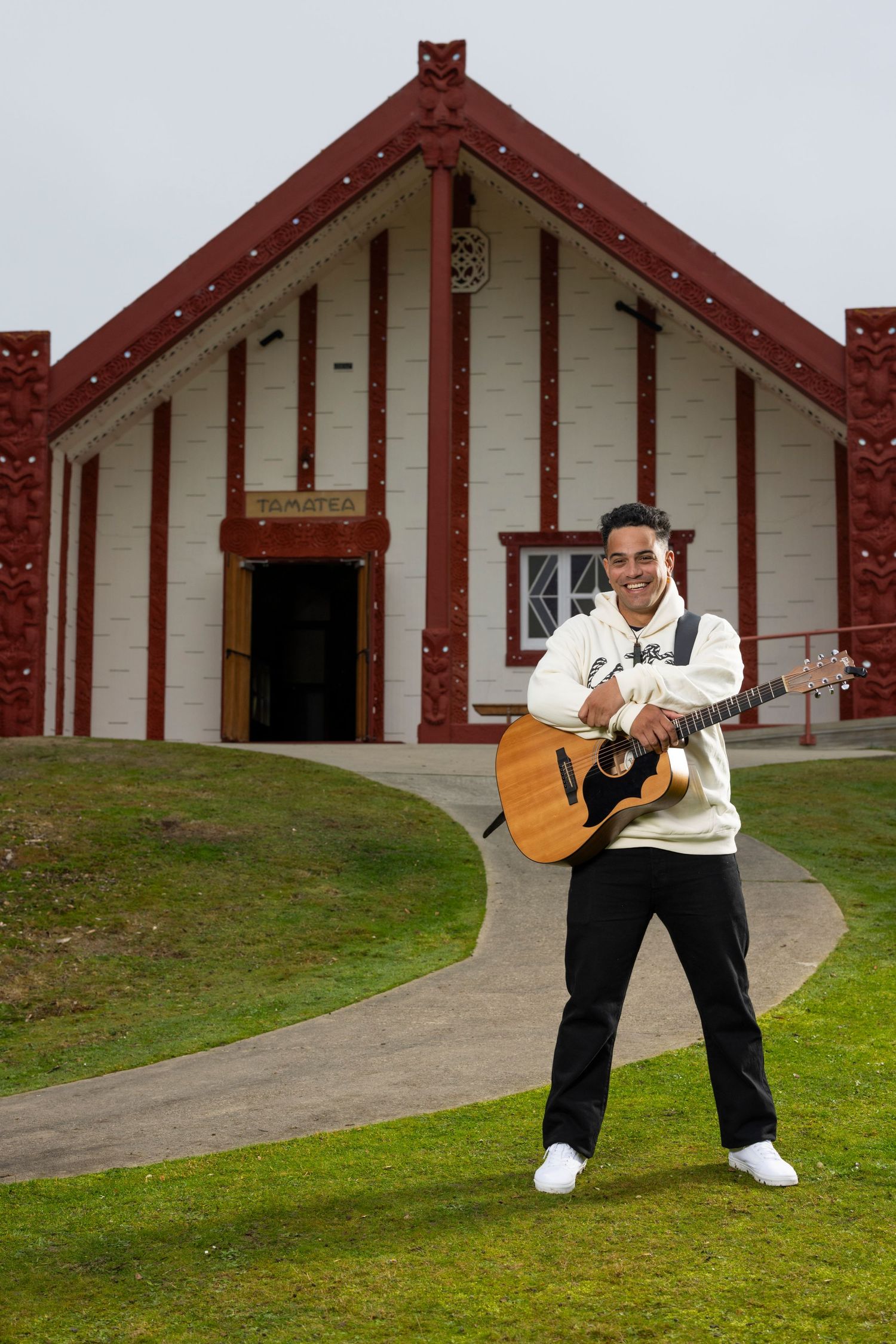 Musician playing acoustic guitar in front of traditional Maori meeting house.