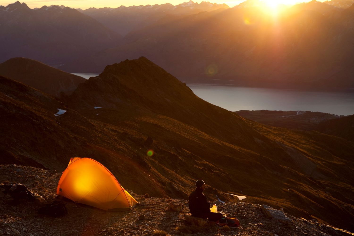 Orange glowing tent illuminates mountainside camp site at dusk with dramatic peaks in background.