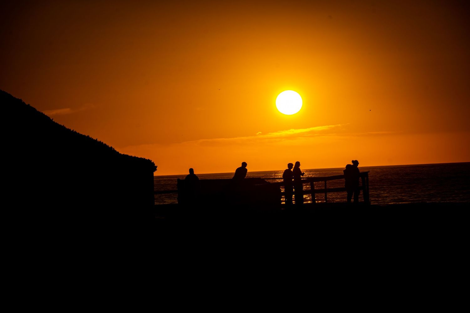 Silhouettes of people watching sunset from observation deck against vibrant orange sky over ocean.