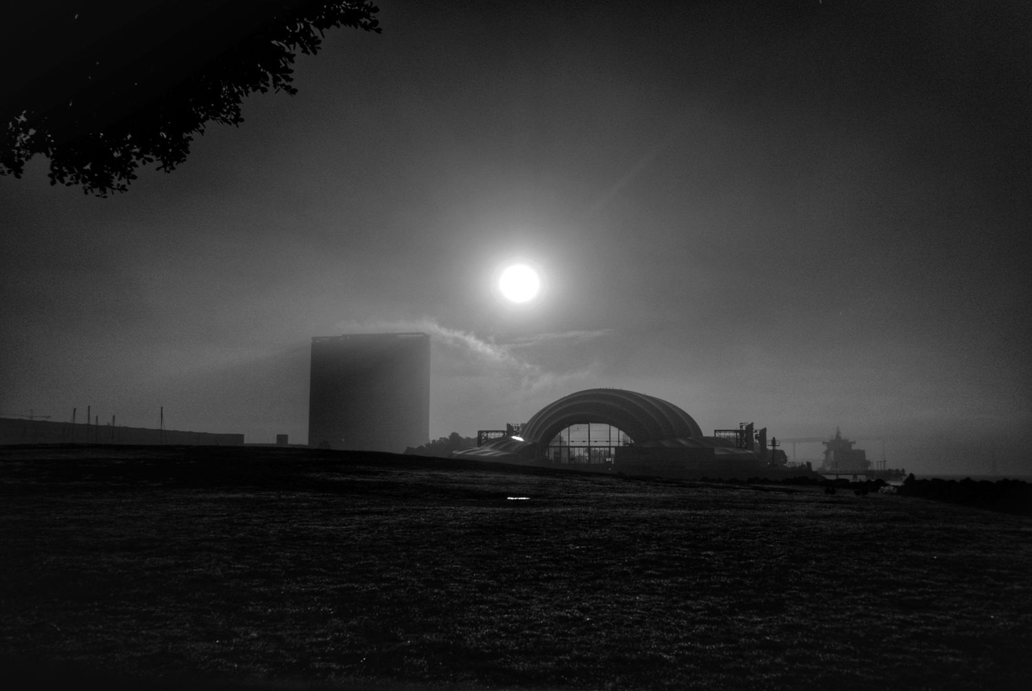 Industrial storage tank and curved building silhouetted against bright sun in black and white.