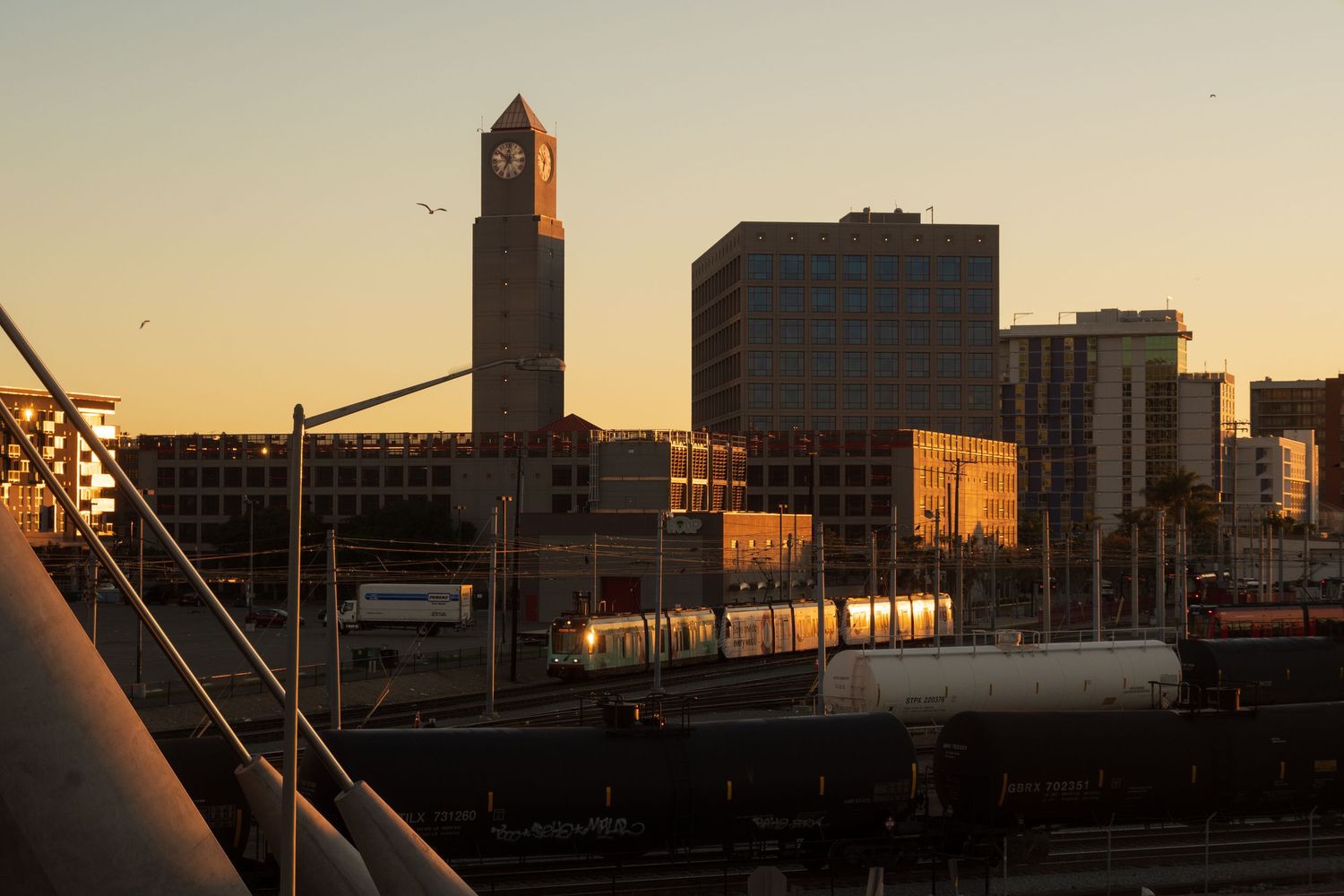 Urban skyline silhouetted against warm sunset sky with industrial buildings and structures.