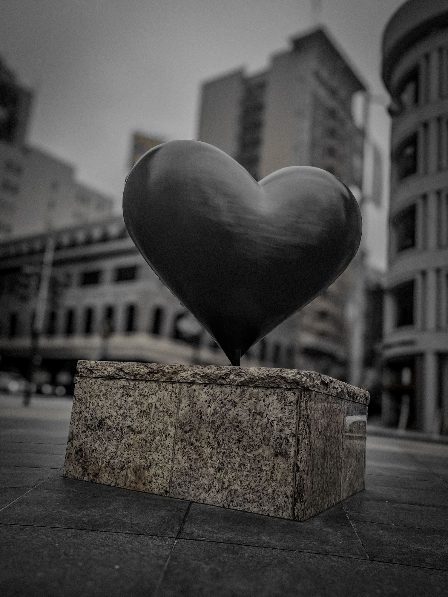 Black metal heart sculpture on concrete pedestal against urban backdrop in black and white.
