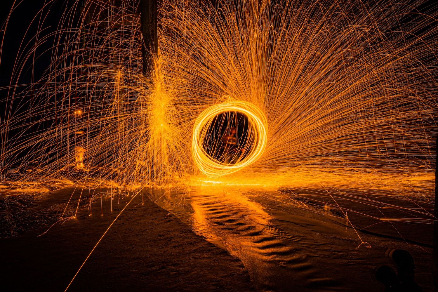 Light painting photography creates glowing circular pattern of sparks reflecting on water surface.