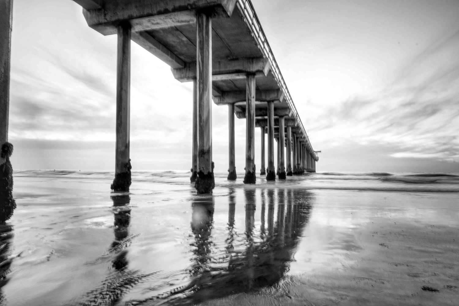 Long exposure of ocean pier stretching into distance with reflective wet sand.