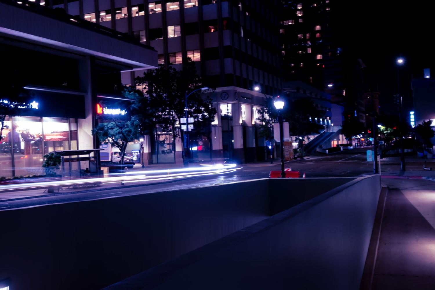 Long exposure night photography captures light trails from traffic on a dark city street with illuminated buildings.