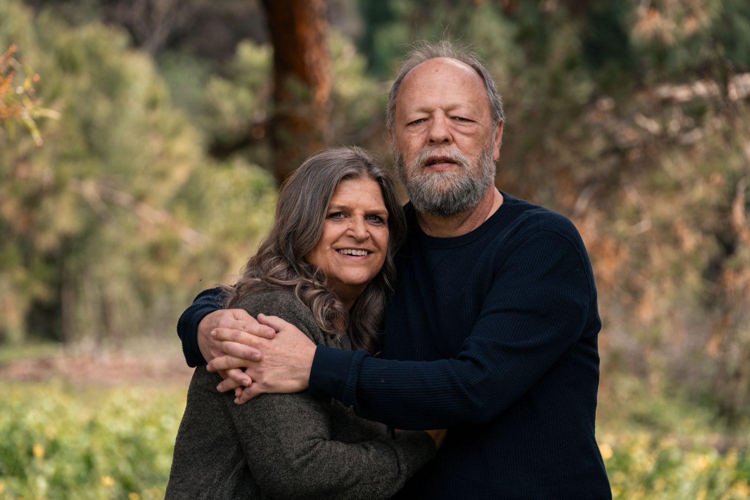 Older couple embracing outdoors, man hugging woman from behind, smiling warmly among green trees.
