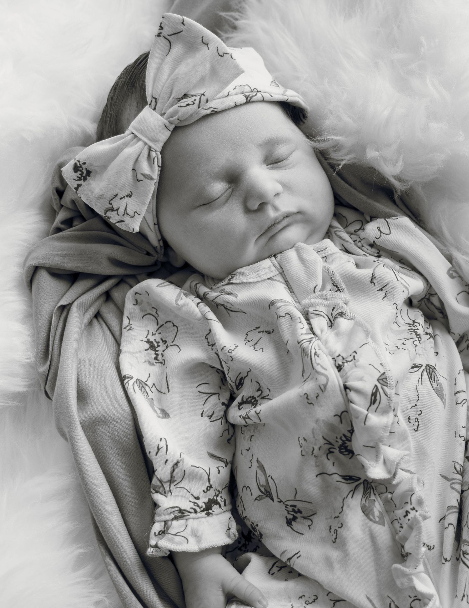 Black and white photo of a sleeping newborn baby girl in a floral outfit and bow headband.