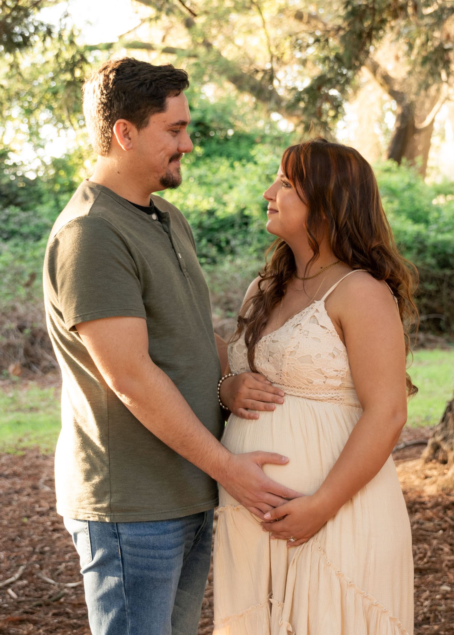 Couple sharing a tender moment outdoors, woman in flowy cream maternity dress cradling baby bump, man in olive shirt, lush green trees.