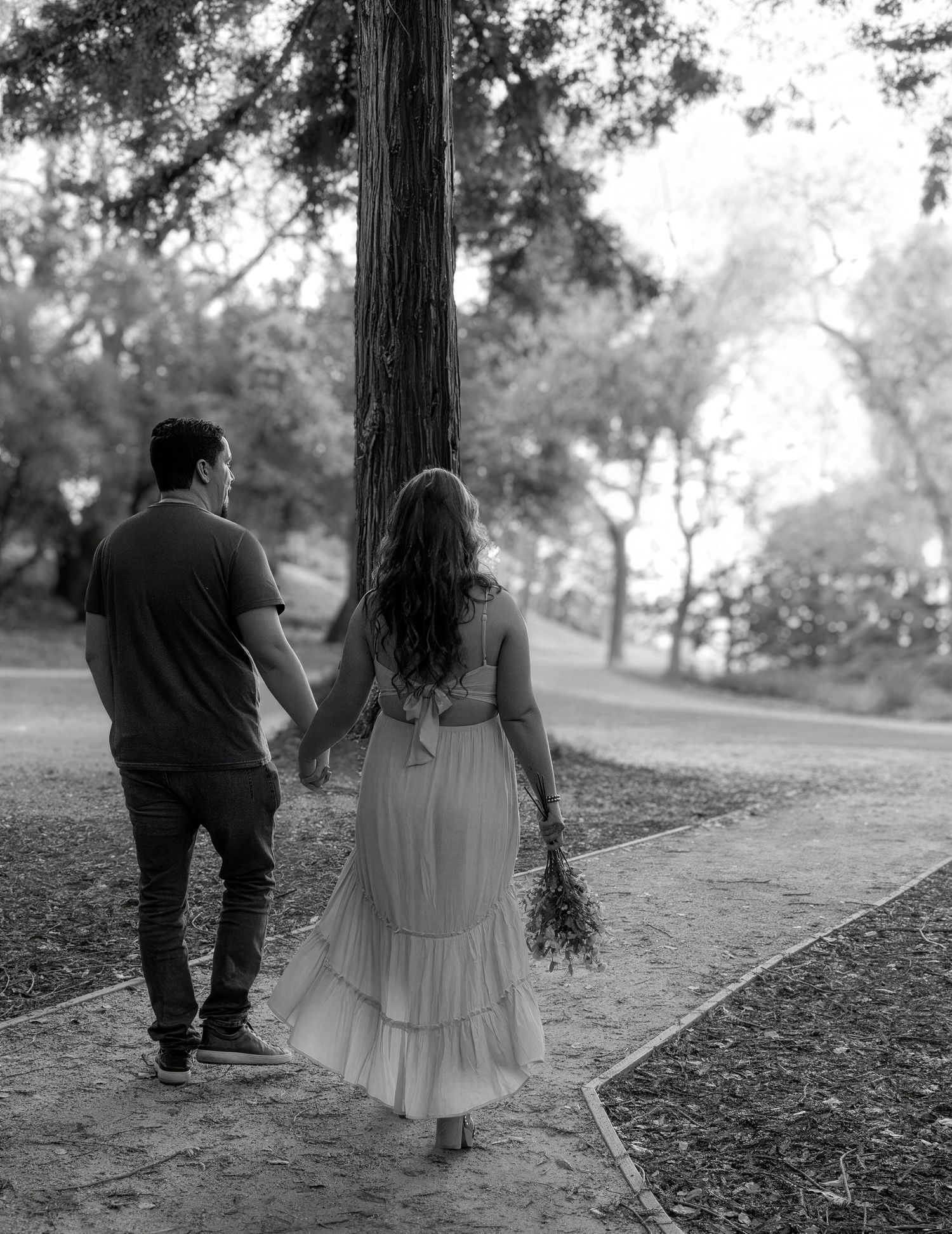 Black and white photo of a couple holding hands walking along a tree-lined path, woman in flowing dress holding wildflowers.