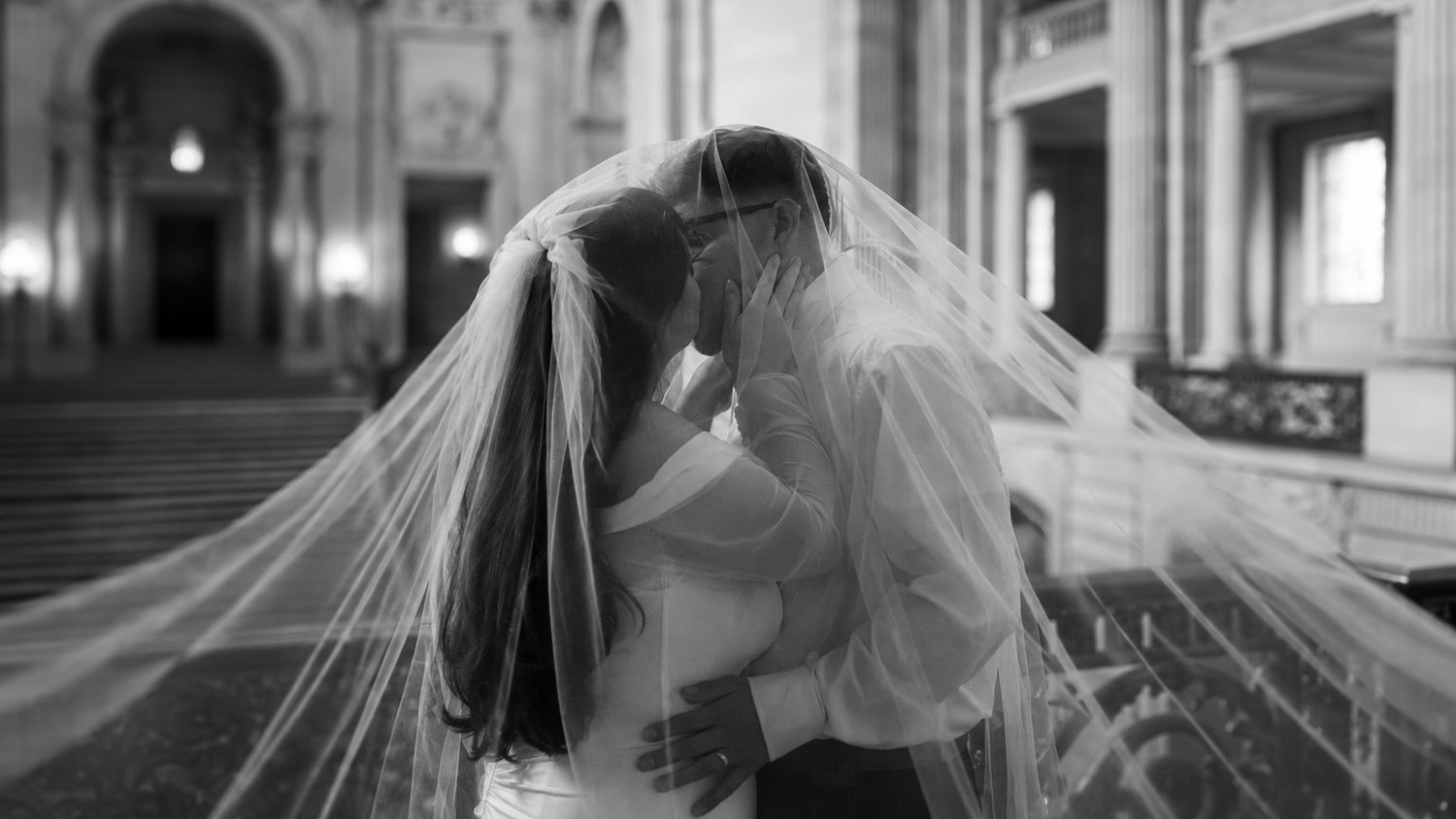 Black and white wedding photo of bride and groom kissing under veil in elegant church.