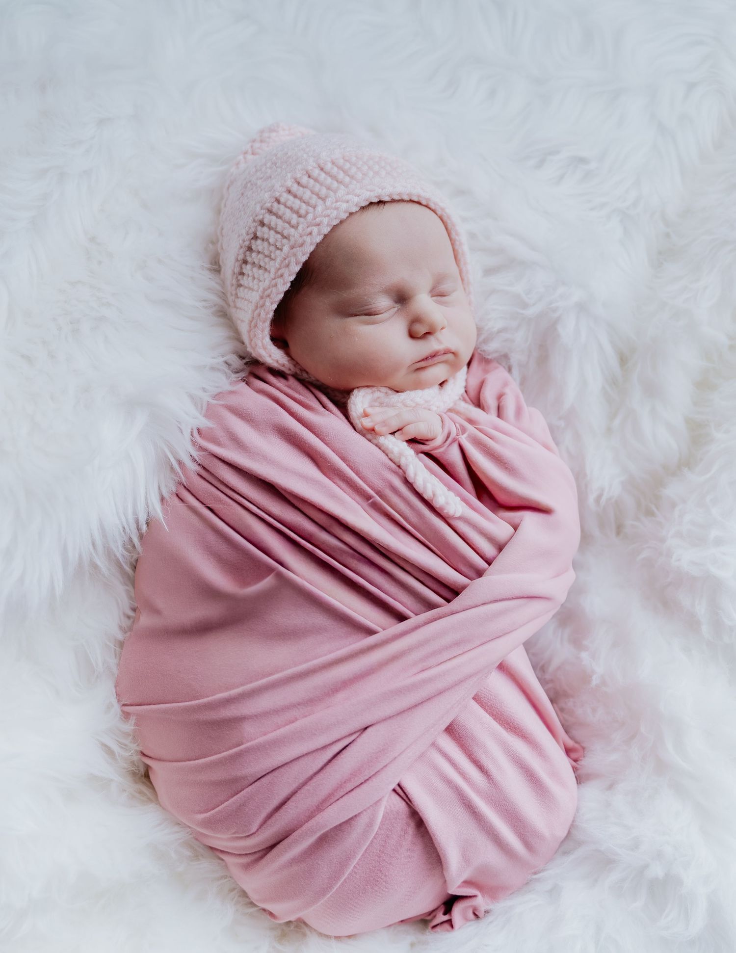 Newborn baby swaddled in pink wrap wearing knit bonnet, sleeping peacefully on white fluffy blanket. Traditional Newborn Photography